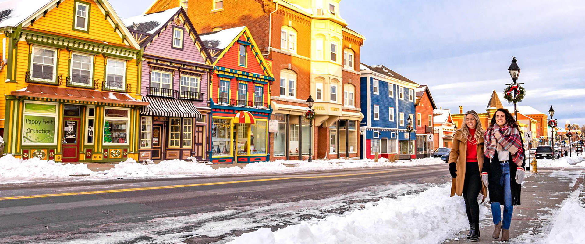 Two women walking along a snow lined street in downtown Yarmouth with colourful storefronts in the background.