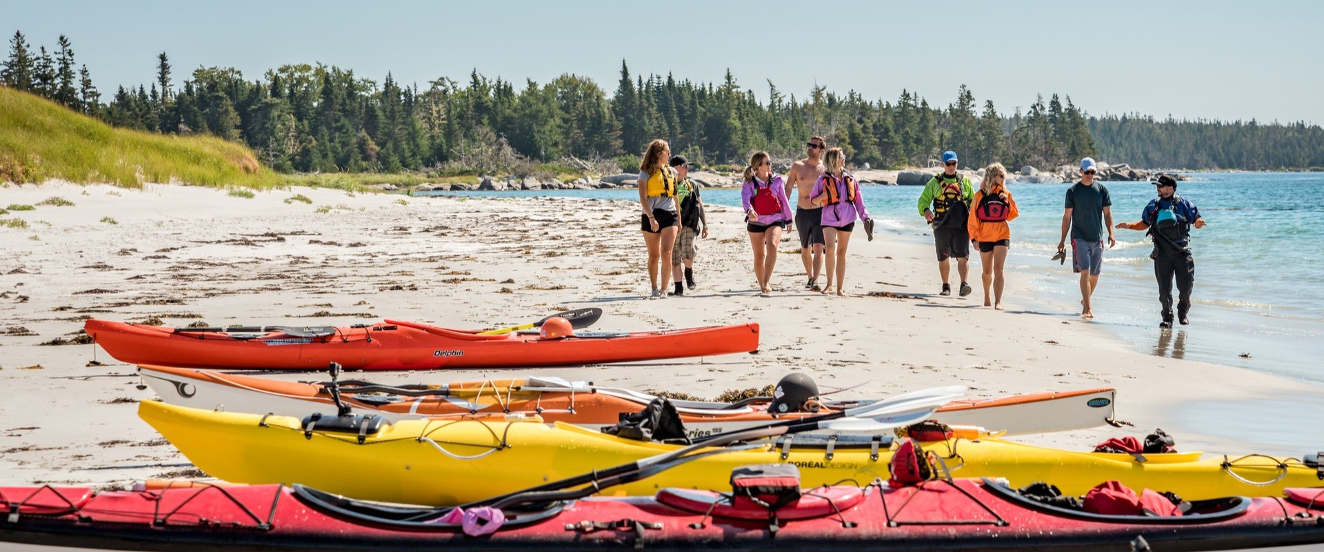 A group of people on a tour walk along a beach towards their kayaks near Kejimkujik National Park Seaside. There are yellow and orange kayaks on the beach in the foreground.