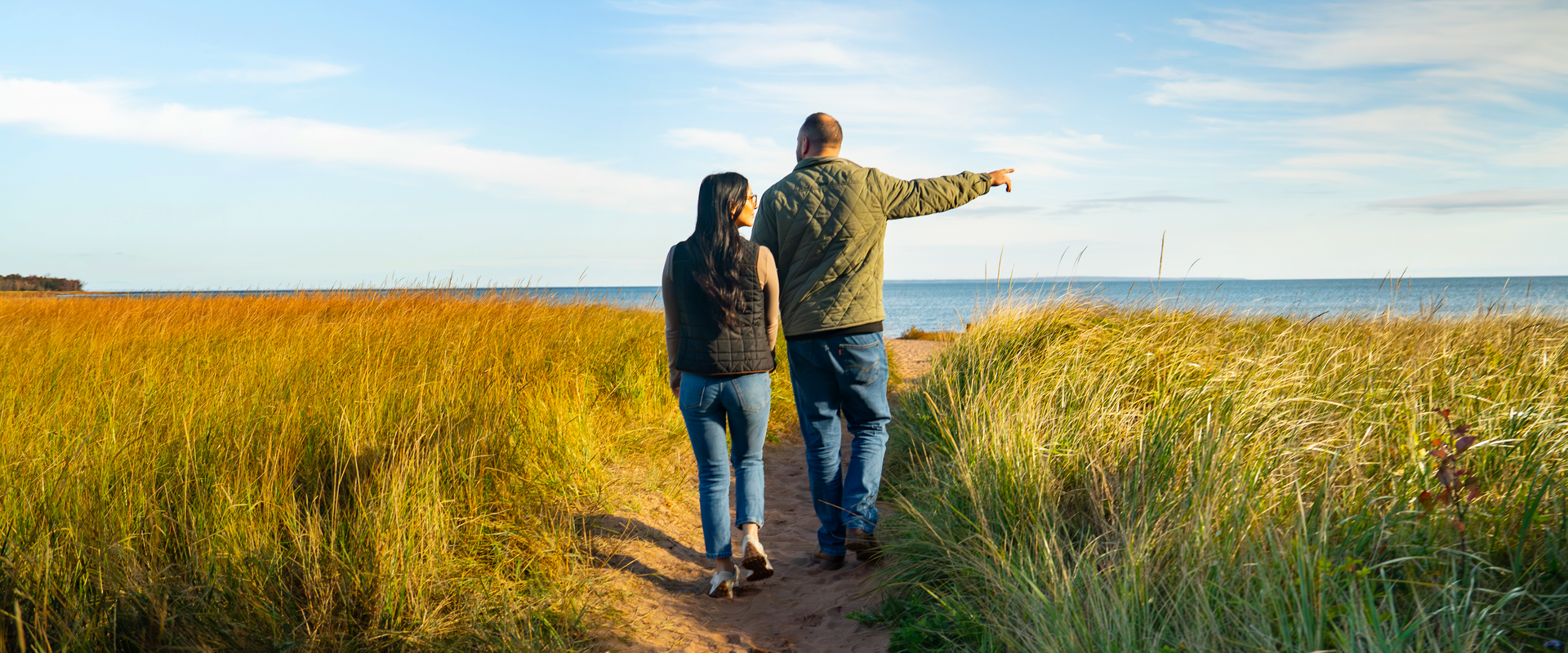 Two people walking over a dune together, one of them is pointing out to sea. 
