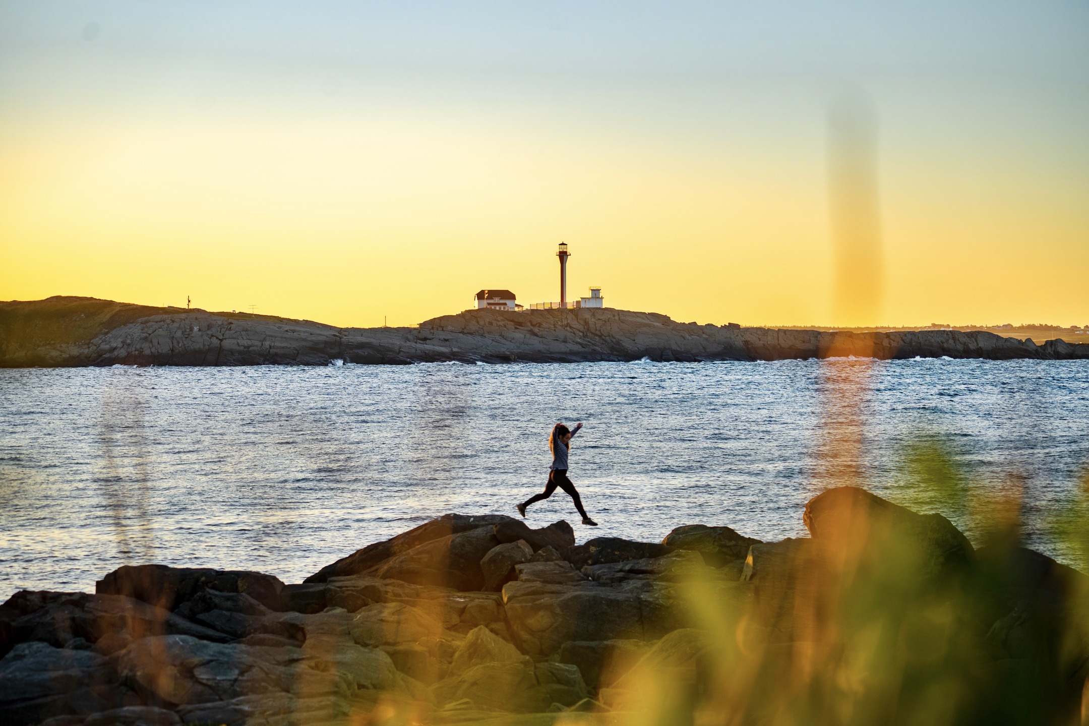 Woman hopping on the rocks along the shore with Cape Forchu Lighthouse in the distance.