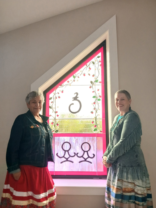 Melinda Robertson and Shannon Monk stand on either side of a commissioned window. 