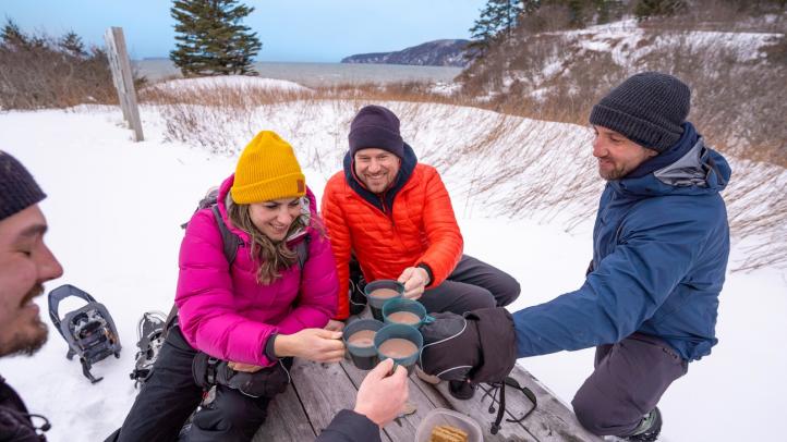 Four people in winter clothing cheers hot chocolate at a picnic table with their snowshoes nearby.