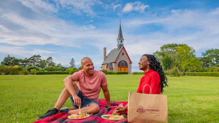 A man and woman sitting on a blanket enjoying a picnic in front of the church at Grand Pre National Historic Site.
