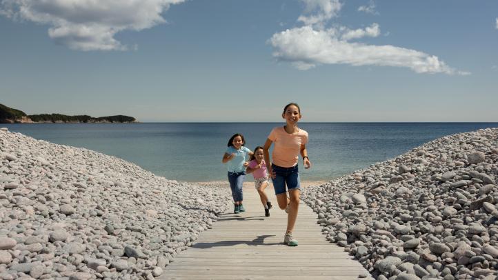 Three kids running on a boardwalk between two piles of rocks with the ocean in the back