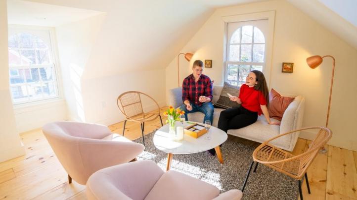 Two people are seated chatting in a bright loft space at August House. It is daytime and the room has four chairs and a small white couch were the two people are chatting. 