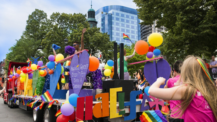 A Pride Parade float in downtown Halifax with pepple on it dancing and people following it. Everyone is dressed in rainbow colours with their clothing and there are rainbow balloons, flags and banners on the parade float. 