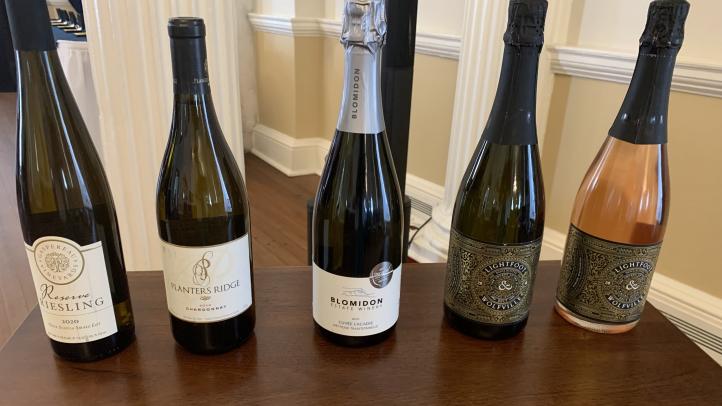 Five bottles of wine on a wooden table. The wines are Gaspereau Vineyards Reserve Reisling, Planters Ridge Chardonnay, Blomidon Estate Winery Cuvee L'acadie, and Lightfoot and Wolfville's Brut and Brut Rose. 