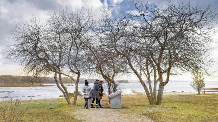 Black Loyalist Heritage Trail in Birchtown. Three people stand in front of a plaque at the end of a trail at the water's edge. There are three leafless trees in the background and the sky is grey. 