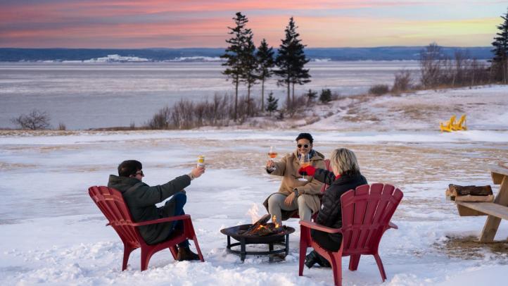 Three people cheers while sitting in red Adirondack chairs in front of a fire outside in a snow covered field.