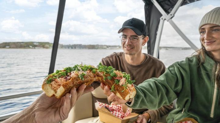 Three people on a boat on the water in Chester. They are 'cheersing' lobster rolls. 