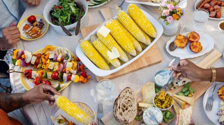 A table top full of food including corn, bread, and kabobs