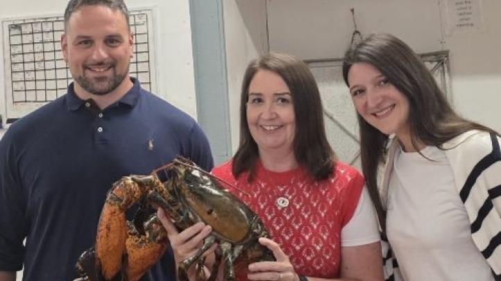 Three people standing together at the Halls Harbour lobster pound. The person in the middle is holding a giant lobster