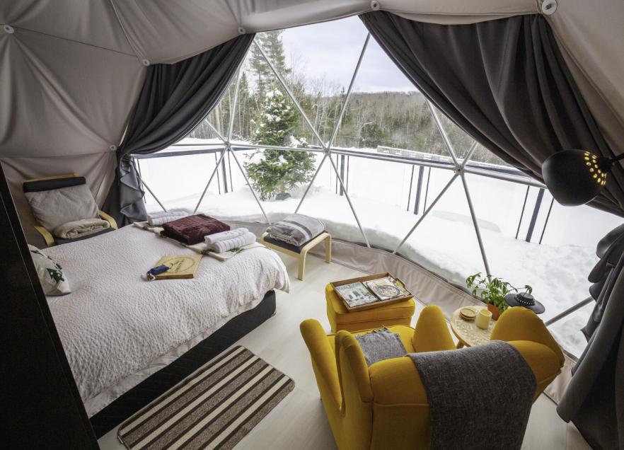 Inside view of camping dome at Valley Sky Luxury Camping. A bed and yellow arm chair face a window looking out at the Annapolis Valley in winter.