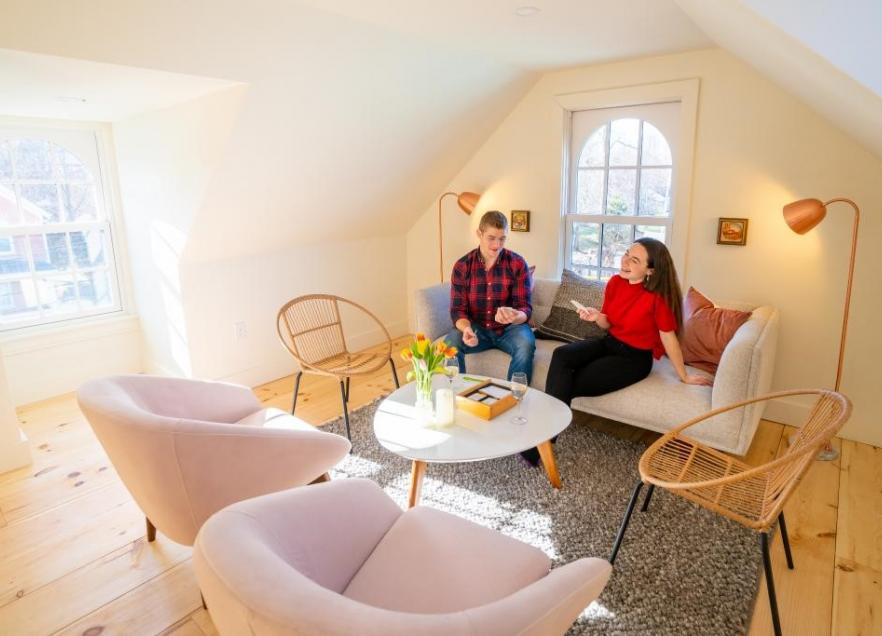 Two people are seated chatting in a bright loft space at August House. It is daytime and the room has four chairs and a small white couch were the two people are chatting. 