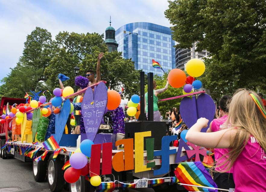 A Pride Parade float in downtown Halifax with pepple on it dancing and people following it. Everyone is dressed in rainbow colours with their clothing and there are rainbow balloons, flags and banners on the parade float. 