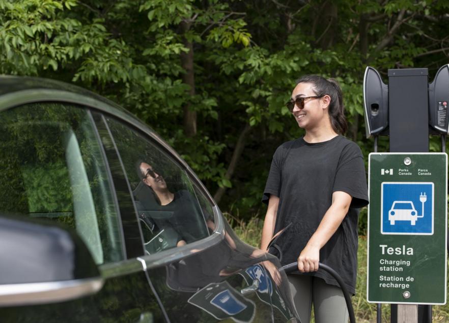 A person is standing next to their EV and hooking it up to a charging station. 