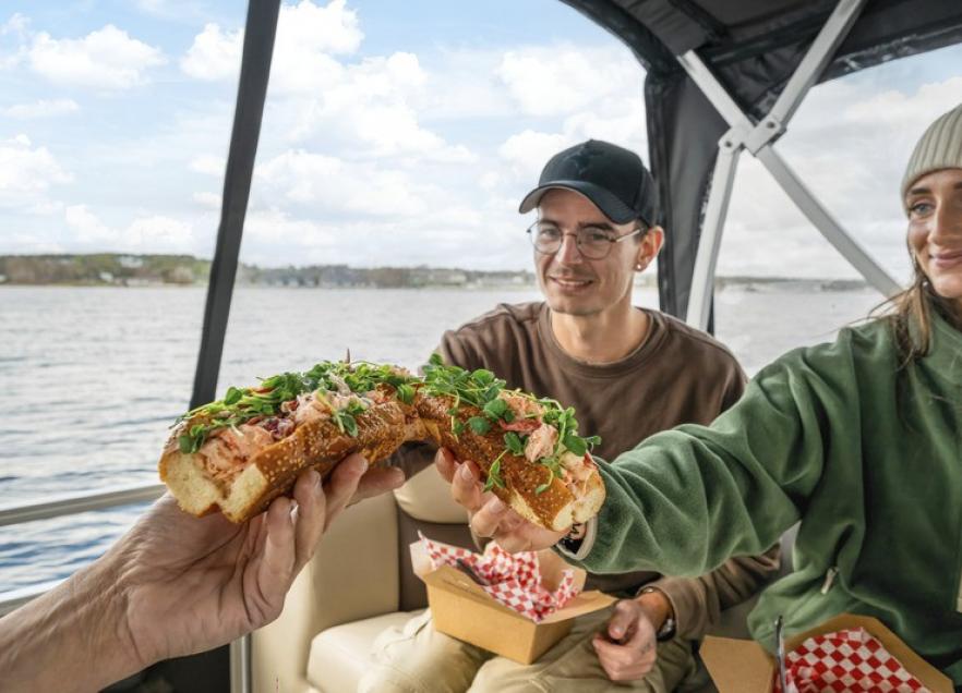 Three people on a boat on the water in Chester. They are 'cheersing' lobster rolls. 