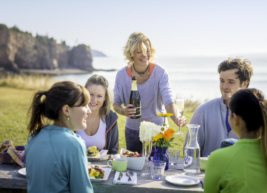 A person serving wine to a group of four people seated at a table on the shore. 