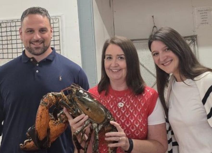 Three people standing together at the Halls Harbour lobster pound. The person in the middle is holding a giant lobster